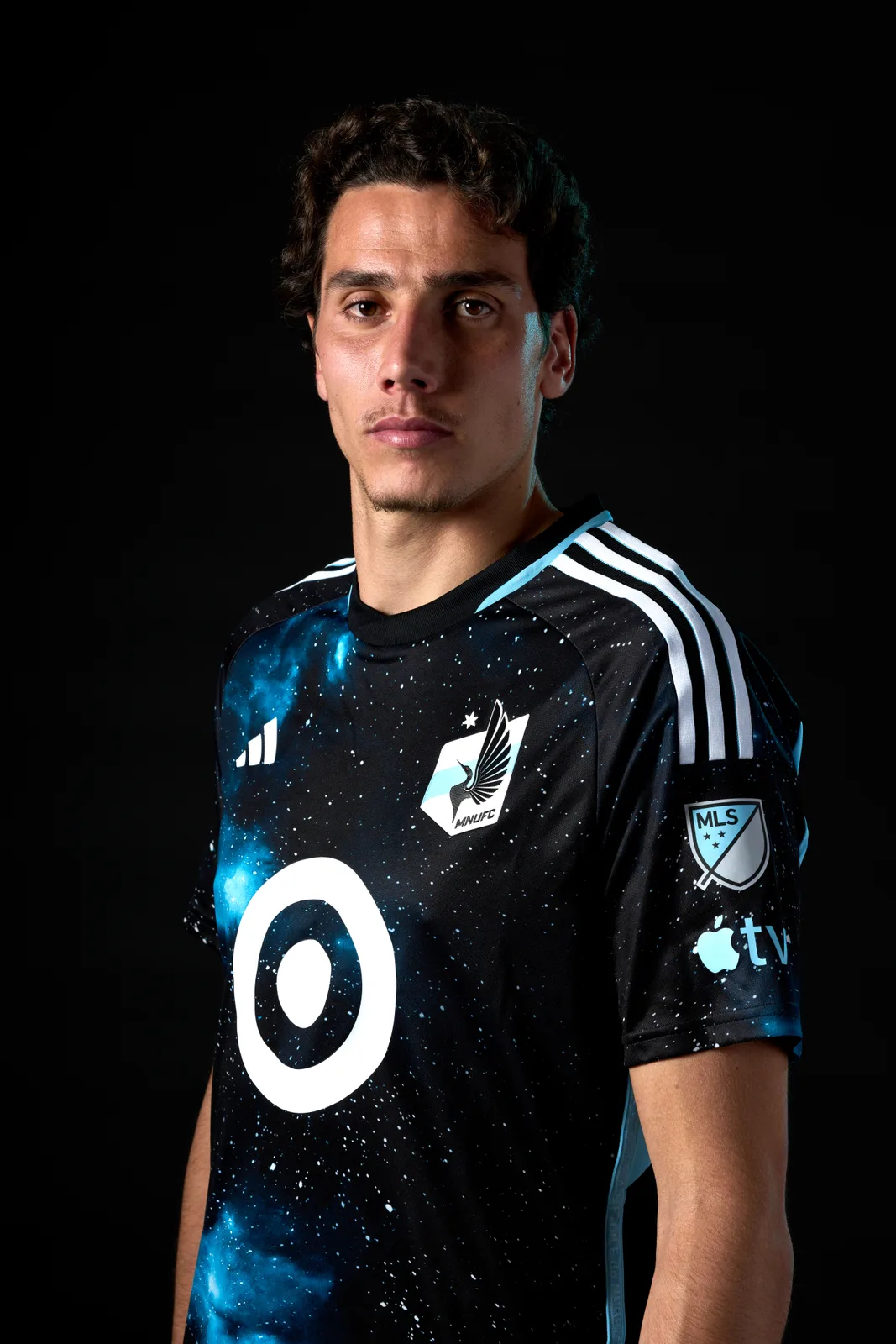 Hugo Bacharach poses against a black background wearing a Minnesota United FC shirt with a starry black and light blue pattern and the MLS badge on the sleeve.