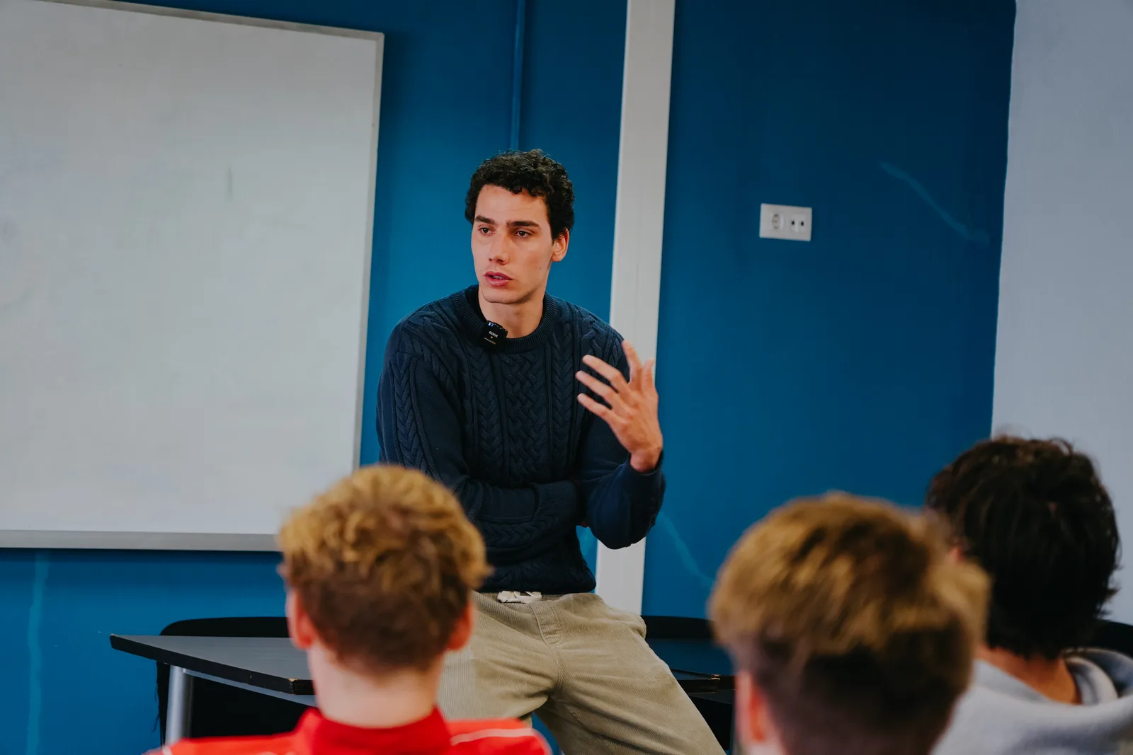Hugo Bacharach, wearing a navy cable-knit sweater and a clip-on microphone, sits on a desk and speaks to a group of young players in a classroom with blue walls and a whiteboard.