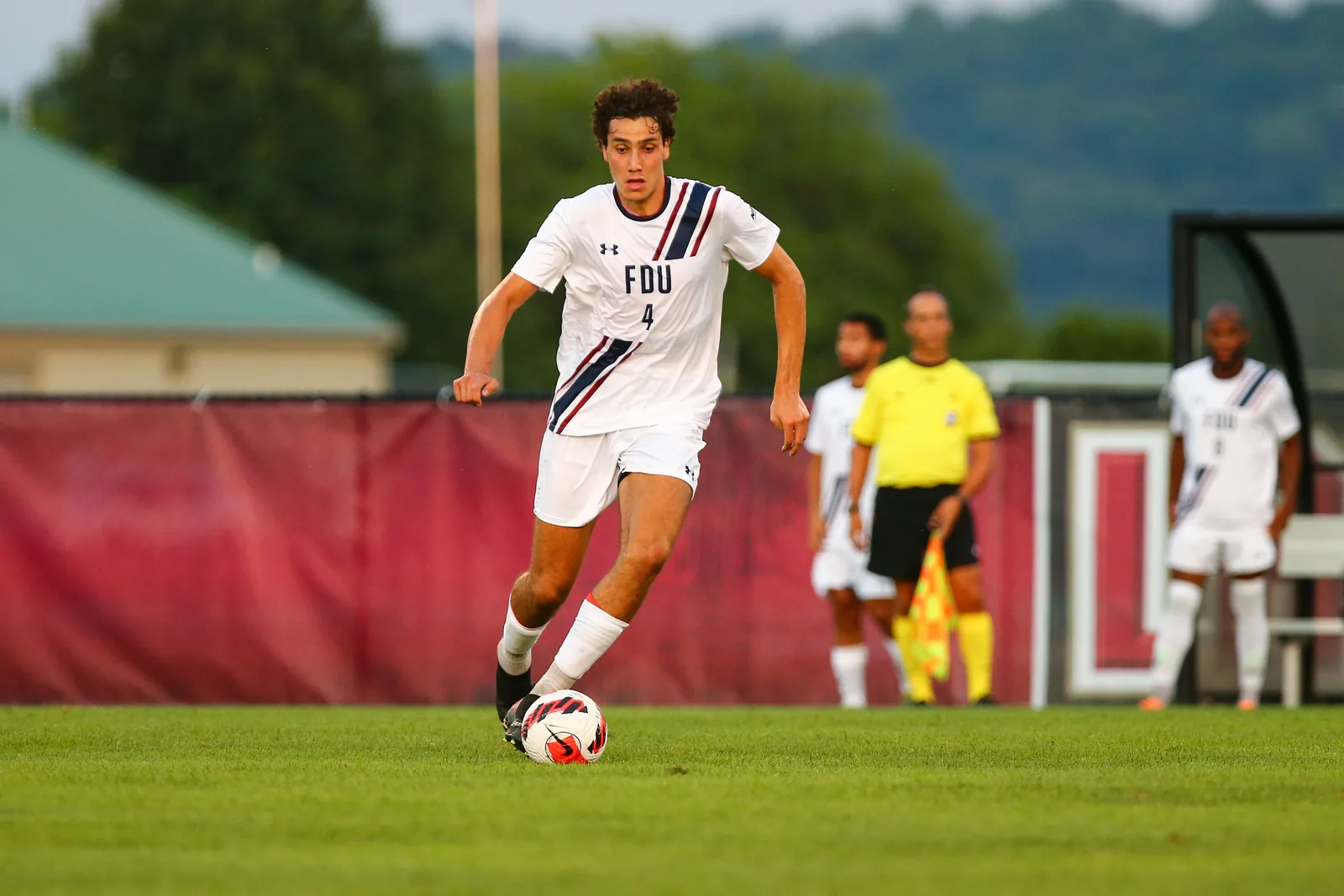 Hugo Bacharach dribbles the ball across the pitch in a white FDU number 4 kit with navy and red stripes during a match.