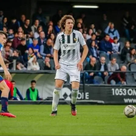 Hugo Bacharach, in a white and black striped kit with white shorts, stands on the pitch during a match as a ball rolls nearby and spectators watch from the stands.