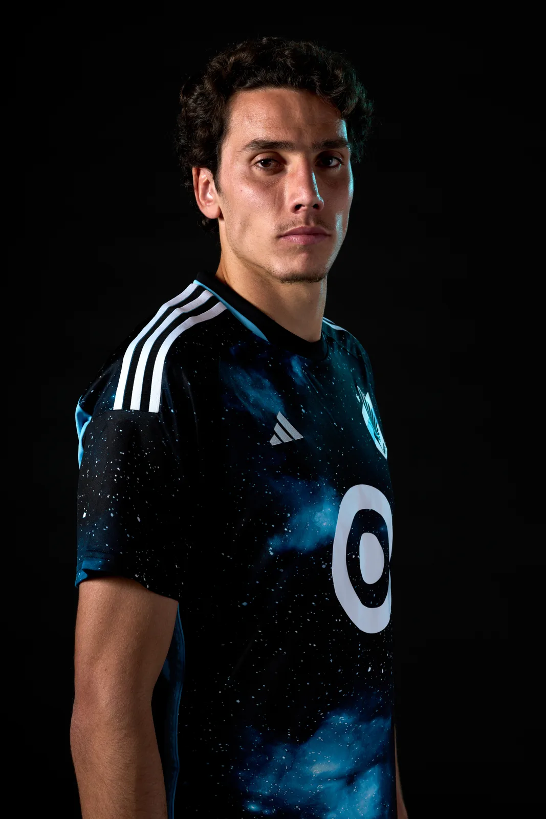Hugo Bacharach poses against a black background in a dark blue starry Minnesota United FC shirt with a white circular sponsor logo.
