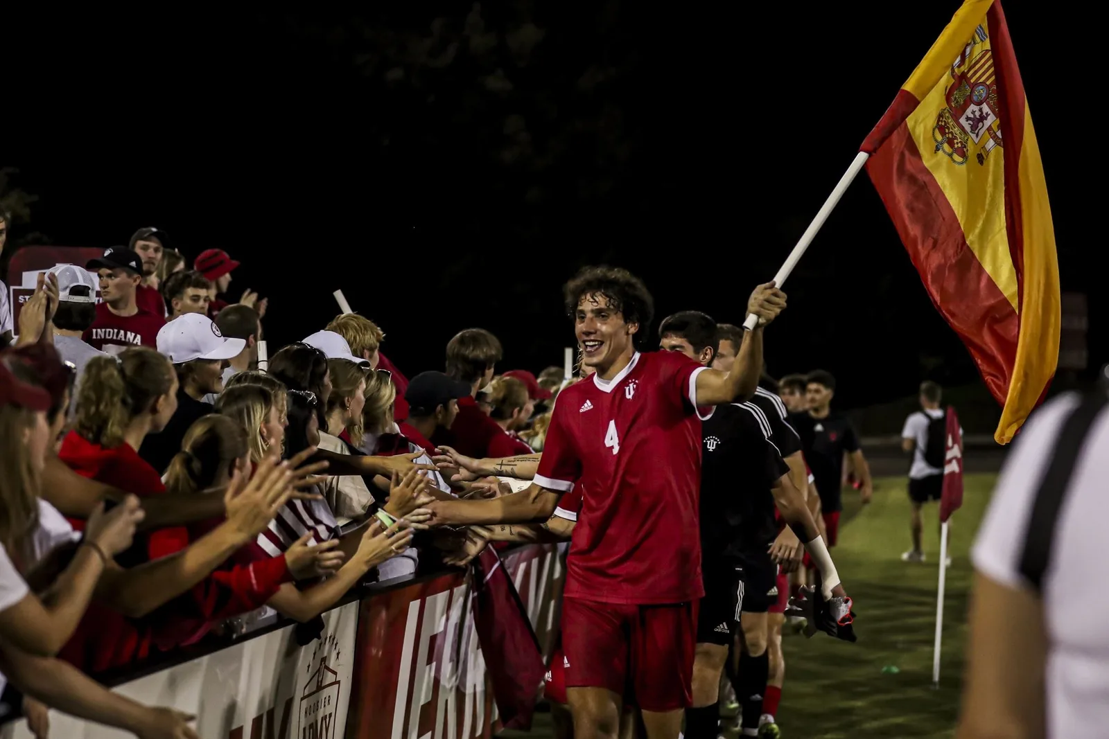 Hugo Bacharach, wearing a red Indiana number 4 kit, holds a Spanish flag and high-fives fans along the barrier at night.