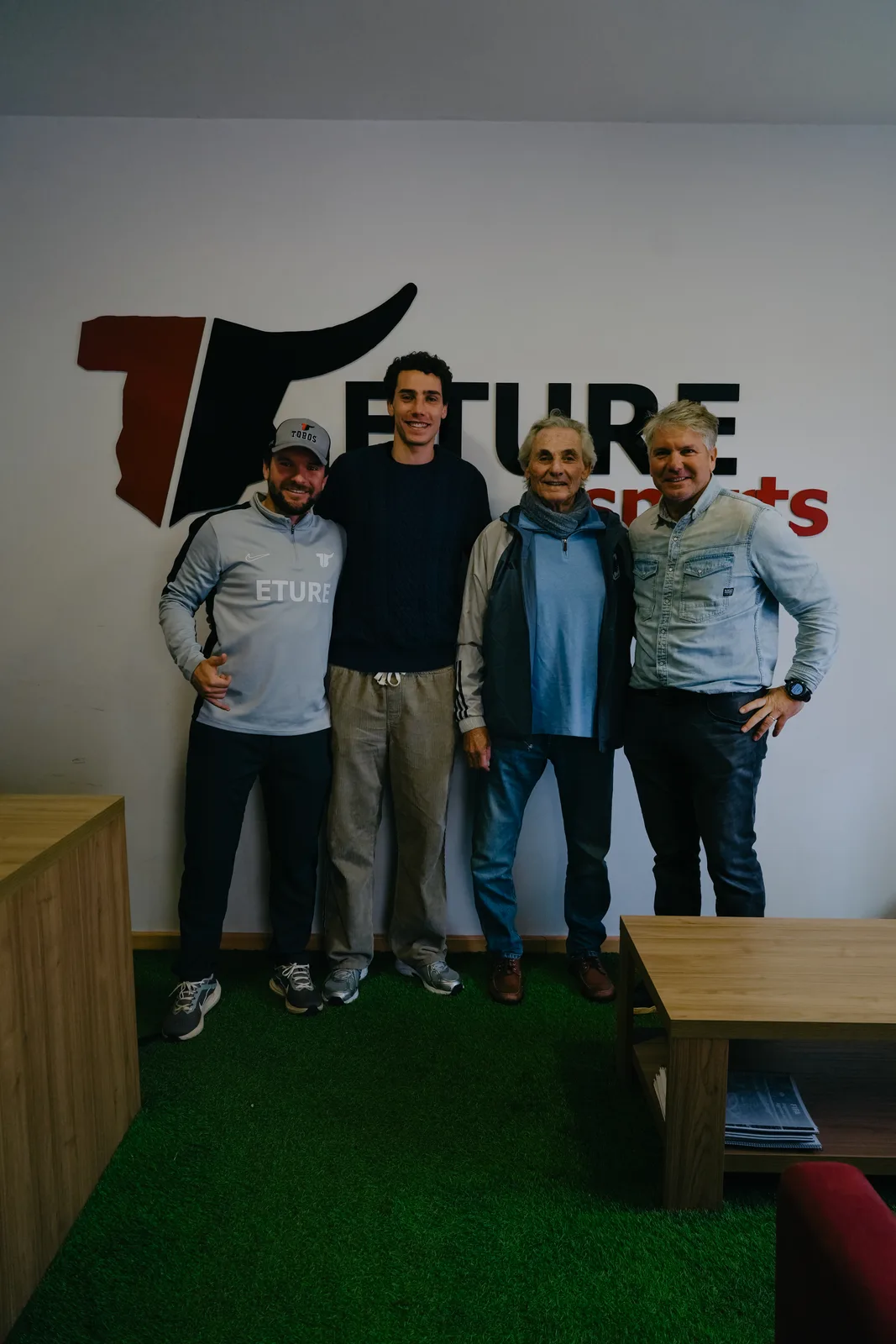 Hugo Bacharach stands with three men in front of the Eture Sports bull logo on a white wall in an office with green artificial turf flooring.