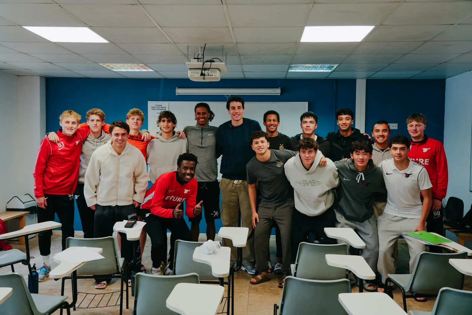 Hugo Bacharach stands with a group of players, several in red Eture training tops, in a classroom with desk chairs and a whiteboard.