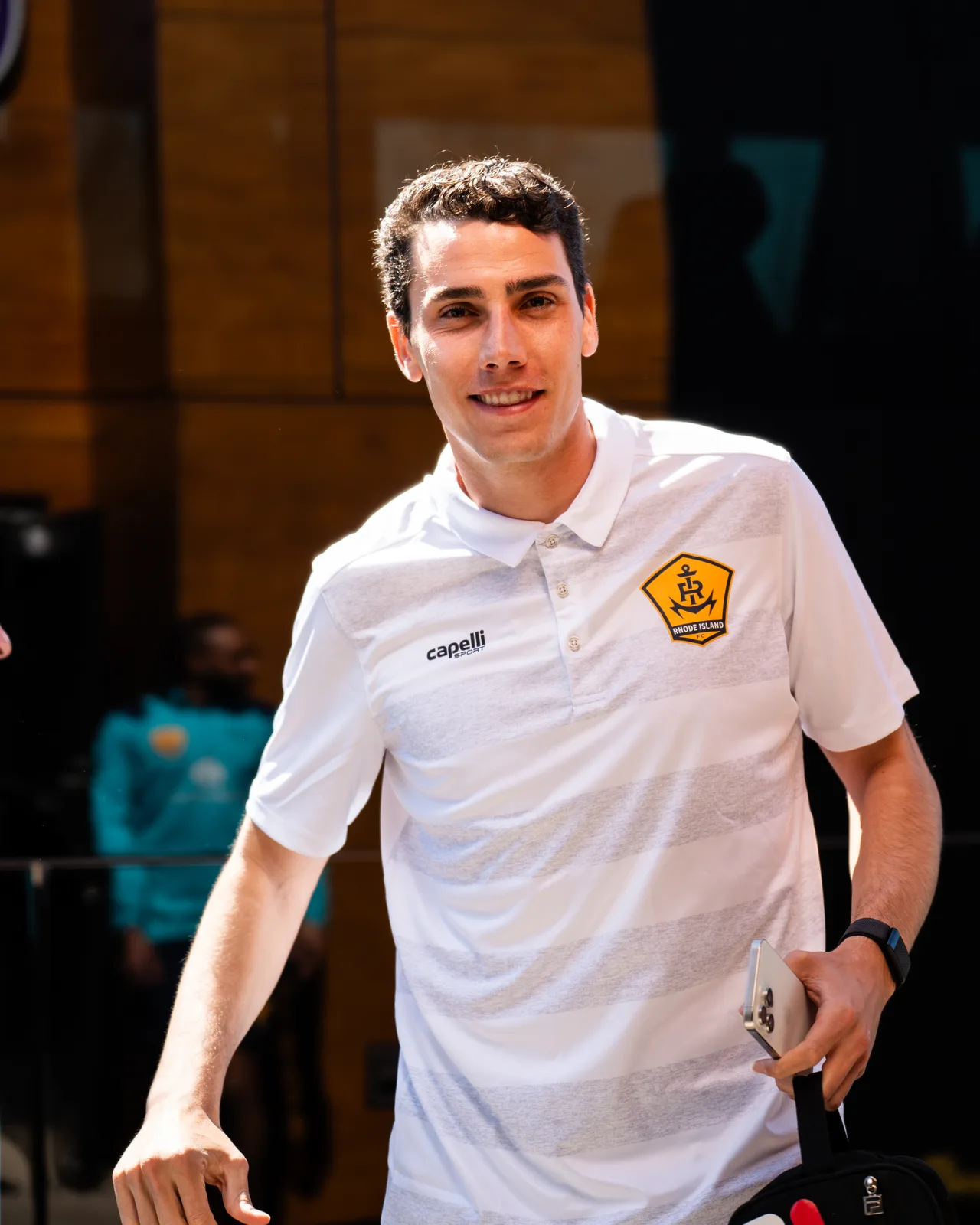 Hugo Bacharach smiles while walking outdoors in a white Rhode Island FC polo shirt, holding a phone and a bag.