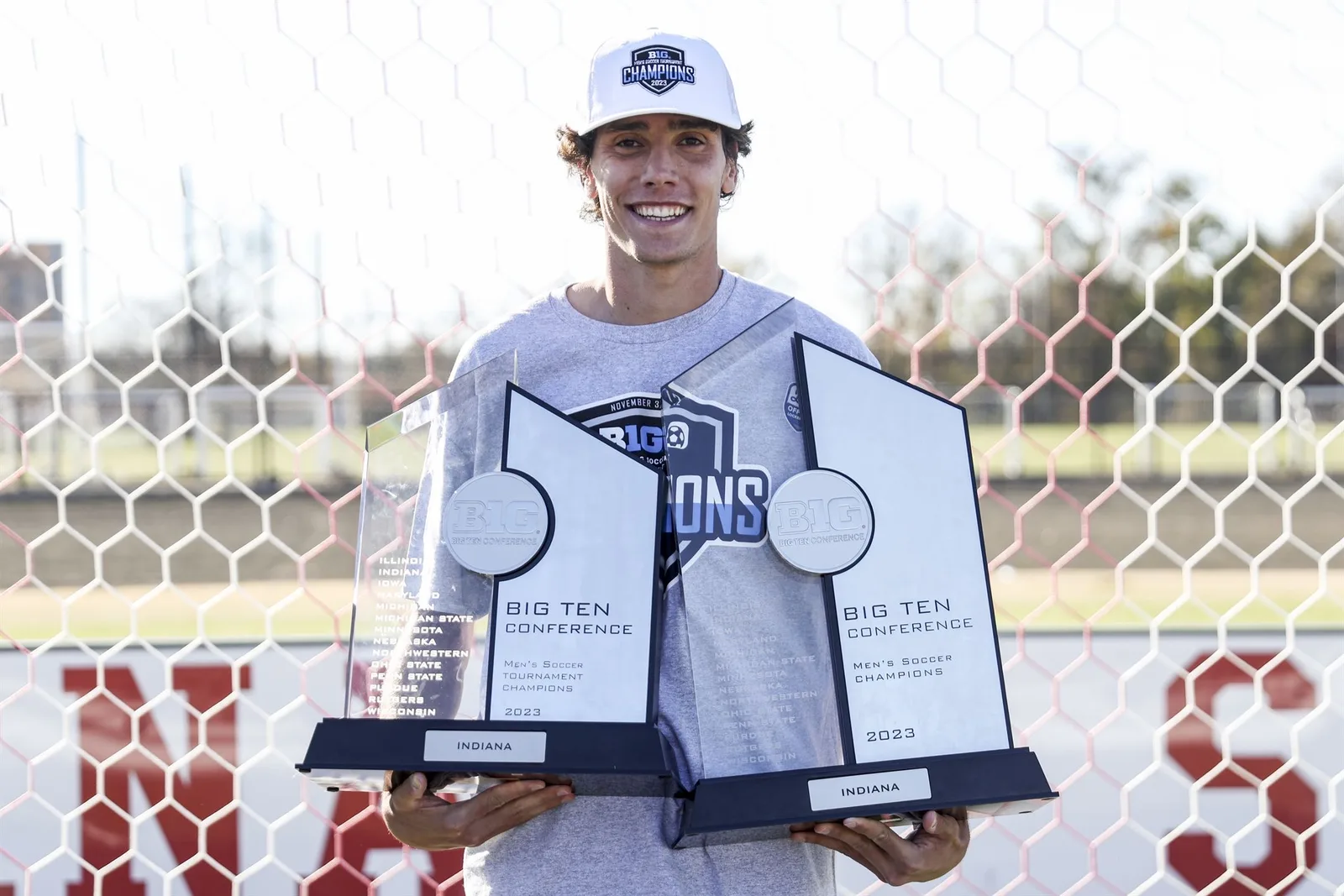 Hugo Bacharach stands in front of a goal net wearing a white Big Ten champions cap and grey champions T-shirt, holding two 2023 Big Ten Conference men's soccer trophies labelled Indiana.
