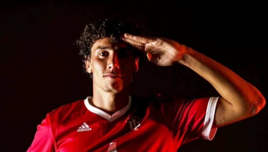 Hugo Bacharach salutes the camera in a red Indiana University Adidas jersey against a dark background.