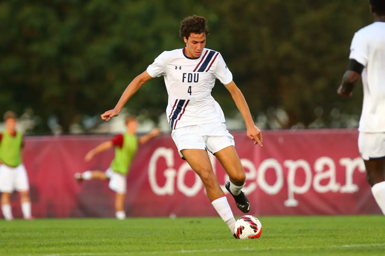 Hugo Bacharach, wearing a white FDU number 4 kit with navy and red stripes, dribbles the ball on a grass pitch during a match.