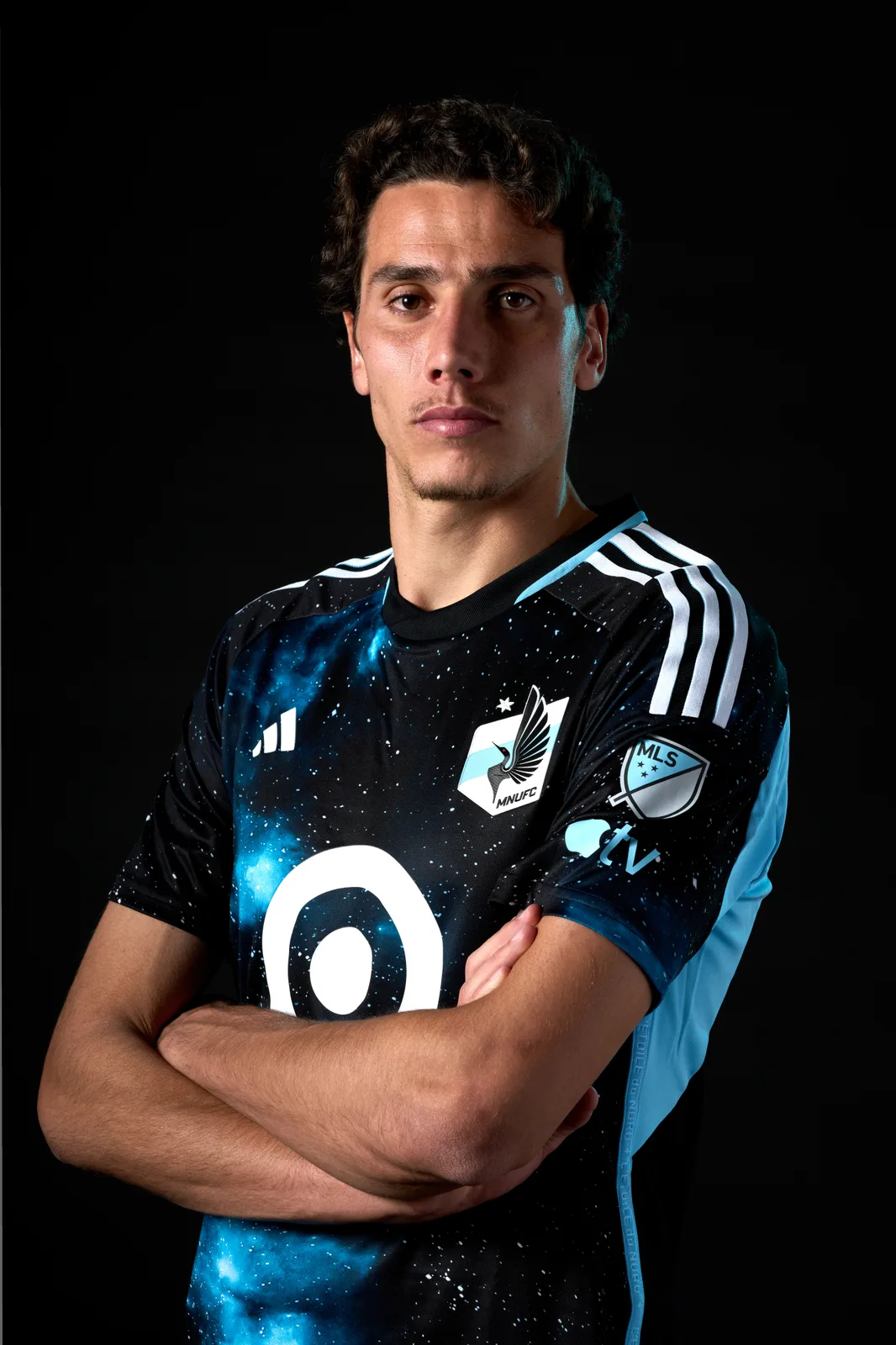 Hugo Bacharach stands with his arms crossed against a black background, wearing a dark Minnesota United FC shirt with a blue starry pattern and the MLS badge on the sleeve.