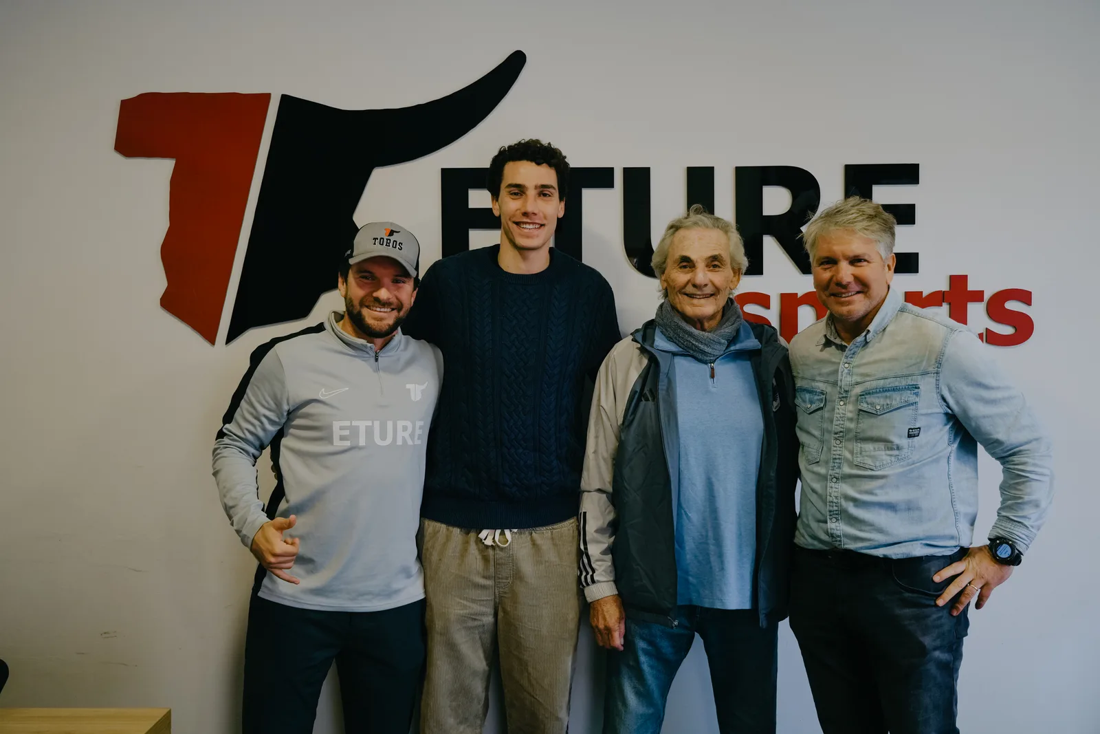 Hugo Bacharach, in a navy cable-knit sweater, stands with three men in front of the Eture Sports logo on a white wall.