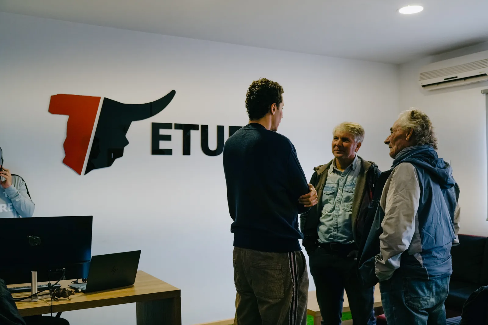 Hugo Bacharach, seen from behind in a navy sweater, talks with two men in the Eture office beside the red and black Eture bull logo on the wall.