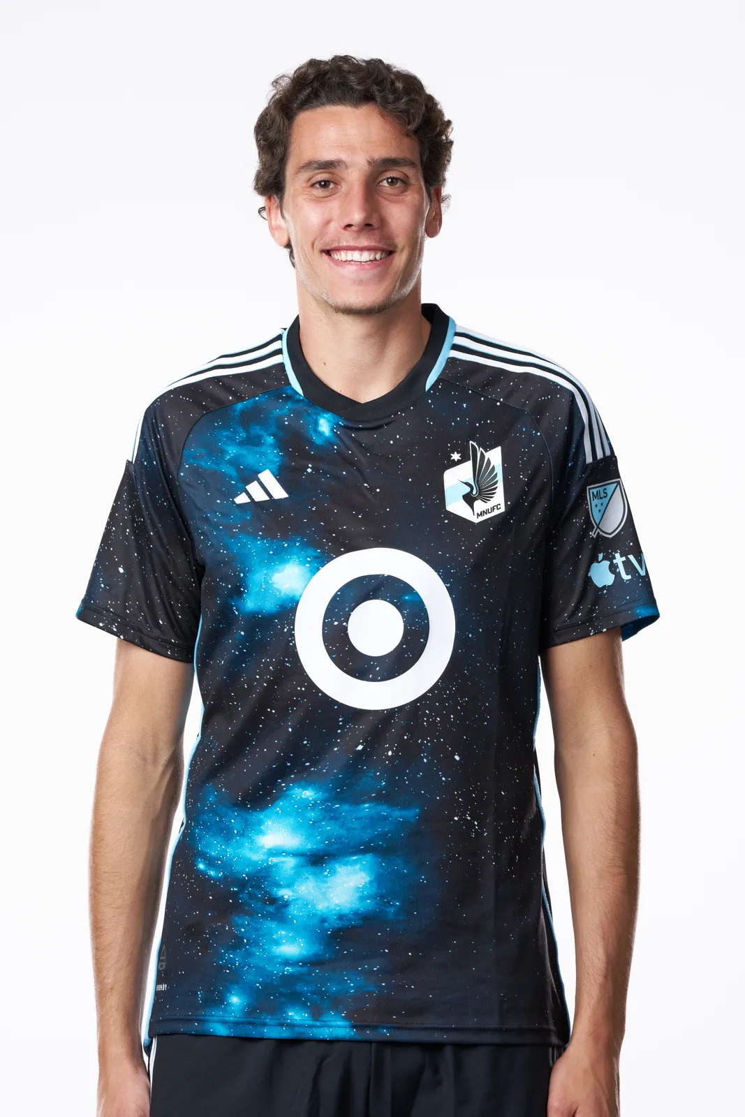Hugo Bacharach stands against a white background in a black and blue galaxy-patterned Minnesota United FC shirt, smiling at the camera.