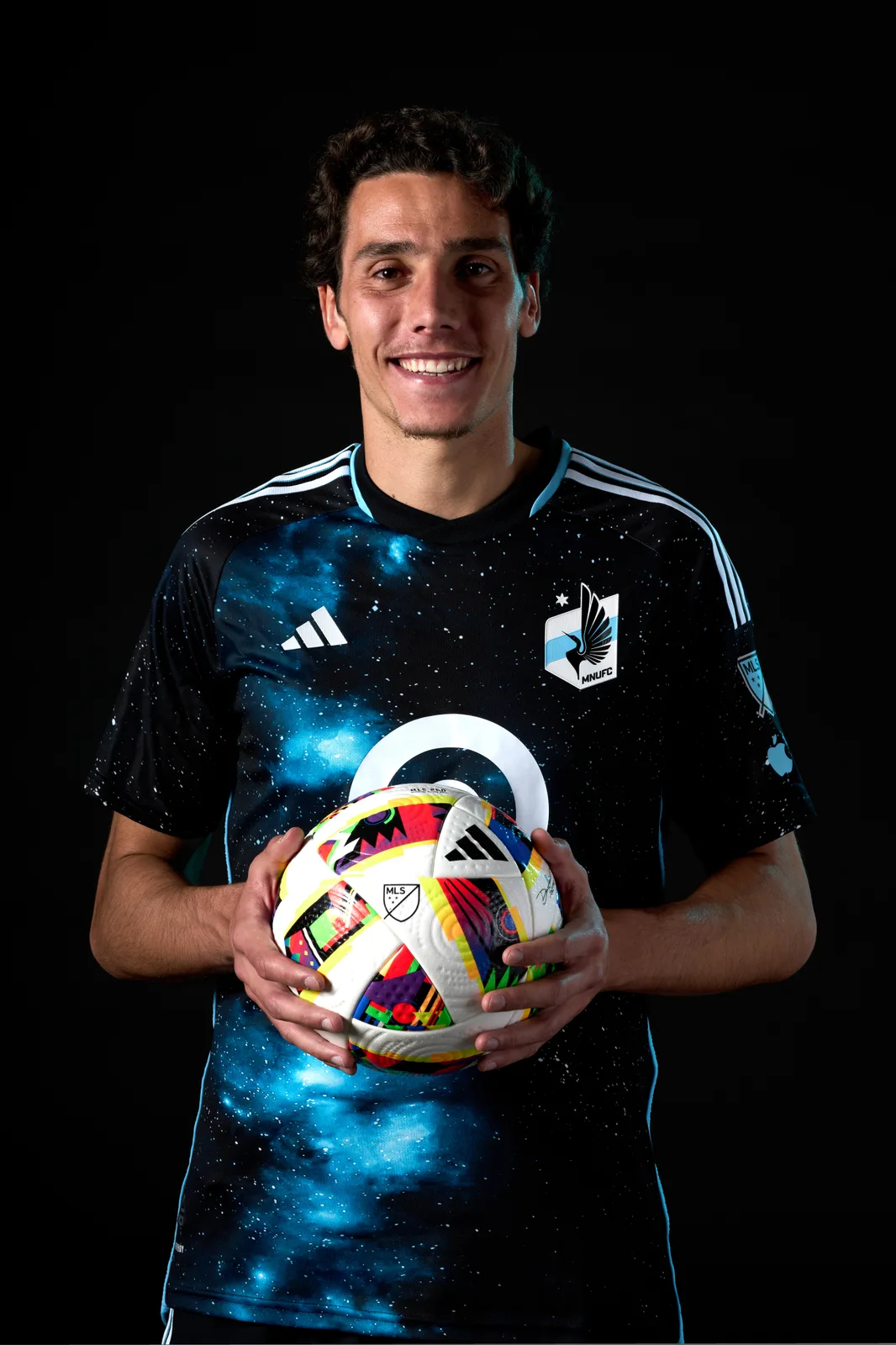 Hugo Bacharach smiles in a dark blue starry Minnesota United FC shirt, holding an MLS ball against a black studio background.