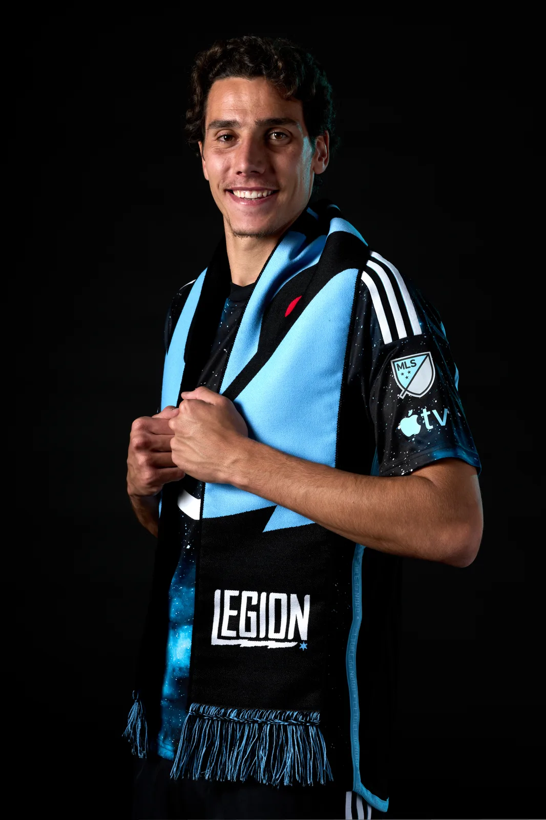 Hugo Bacharach smiles in a studio portrait against a black background, wearing a black and light blue MLS jersey with a matching scarf around his neck.